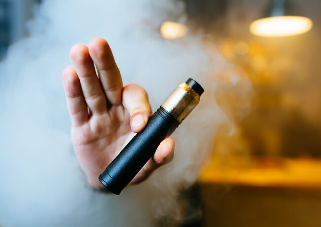 Person, der holder en sort e-cigaret i hånden med damp i baggrunden